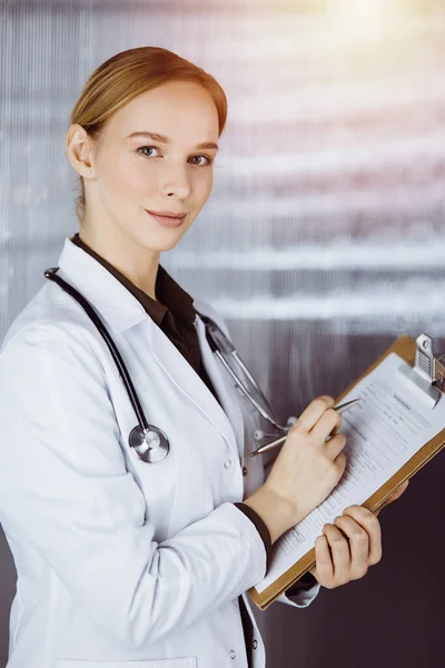 Alegre sorridente médico feminino usando prancheta na clínica. Retrato de mulher médica amigável no trabalho. Serviço médico no hospital. Conceito de medicina — Fotografia de Stock