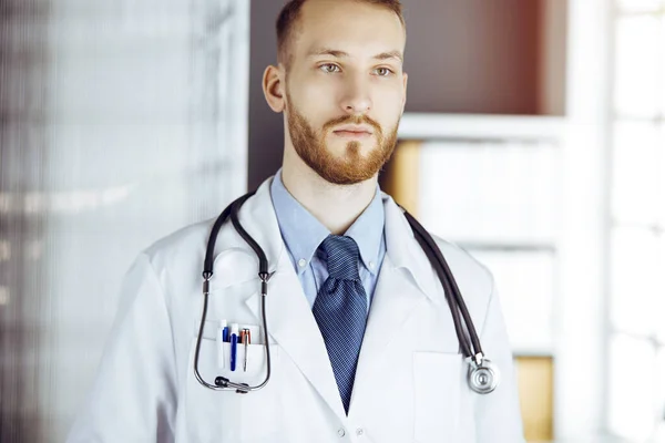 Friendly bearded doctor standing straight in sunny clinic. Portrait of physician. Medicine concept