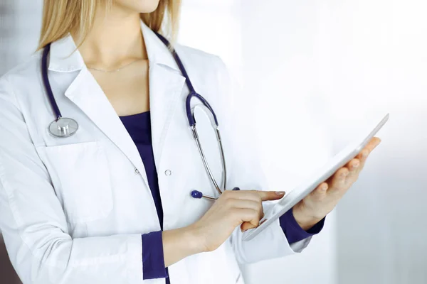 Unknown woman-doctor is holding a tablet computer in her hands, while standing in a sunny clinic cabinet. Female physician at work, close-up. Perfect medical service in a hospital. Medicine concept — Stock Photo, Image