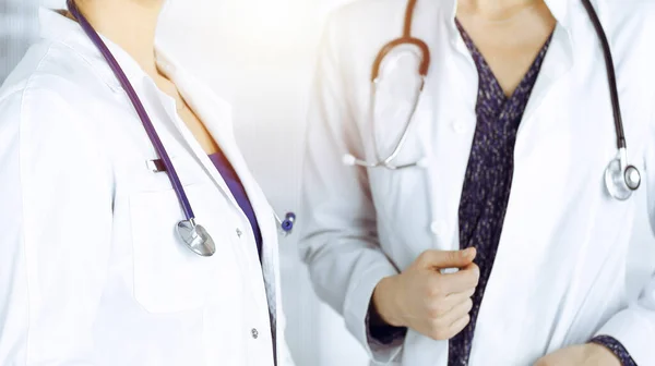 Dos médicas están hablando de terapia médica, mientras están juntas en una clínica soleada. Los médicos usan estetoscopios en el trabajo. Trabajo en equipo en medicina —  Fotos de Stock