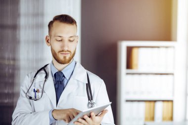 Dost canlısı sakallı doktor iş yerindeki güneşli klinikte tablet bilgisayar kullanıyor. Tıp konsepti