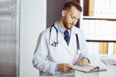 Dost canlısı sakallı doktor güneşli klinikteki panoda oturuyor ve yazıyor. Tıp konsepti