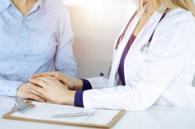 Bilinmeyen kadın doktor, bir klinikteki güneşli dolapta yan yana otururken hastasına güven vermek için elini tutuyor. Kadın hekim bir pano ve steteskop kullanıyor.