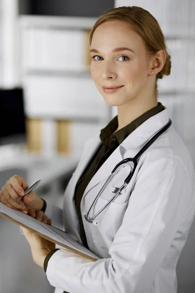 Friendly smiling female doctor using clipboard in clinic. Portrait of friendly physician woman at work. Perfect medical service in hospital