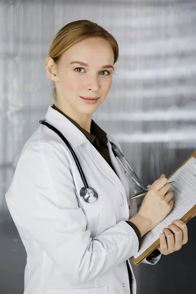 Friendly smiling female doctor using clipboard in clinic. Portrait of friendly physician woman at work. Perfect medical service in hospital