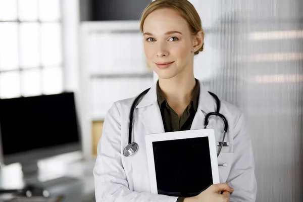 Friendly smiling female doctor using clipboard in clinic. Portrait of friendly physician woman at work place. Perfect medical service in hospital