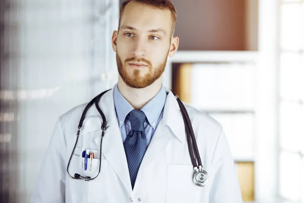 Vriendelijke bebaarde dokter die recht in een zonnige kliniek staat. Portret van een arts. Geneesmiddelenconcept — Stockfoto