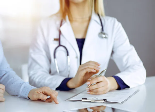 Unknown woman- doctor is listening to her patient, while sitting together at the desk in the sunny cabinet in a clinic. Female physician with a stethoscope is writing at clipboard, close up. Perfect — Stock Photo, Image