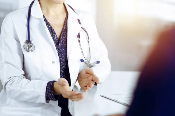 Donna-medico sconosciuto sta parlando con la sua paziente della sua diagnosi, mentre seduti insieme alla scrivania nel gabinetto soleggiato in una clinica. Medico sul posto di lavoro, primo piano. Servizio medico perfetto — Foto Stock