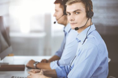 Kulaklıklı iki genç adam güneşli, modern bir ofiste masada oturup müşterileri dinliyor. İş yerindeki çağrı merkezi operatörleri