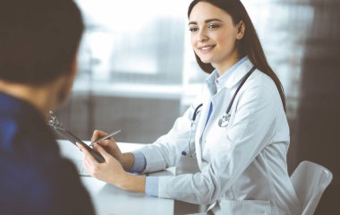 Dost canlısı genç bayan-doktor, birlikte bir dolapta masada otururken, hastasını dinliyor. Hekim elinde ilaç doldurmak için bir dosya tutuyor.