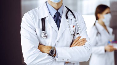 Koruyucu maskeli kimliği belirsiz erkek doktor klinikte kollarını kavuşturmuş duruyor. Coronavirus salgını sırasında ilaç konsepti. Covid 2019