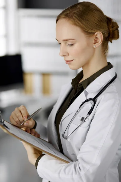 Freundlich lächelnde Ärztin mit Klemmbrett in der Klinik. Porträt einer freundlichen Ärztin bei der Arbeit. Perfekte medizinische Versorgung im Krankenhaus — Stockfoto