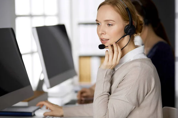 Femme d'affaires amicale parlant par casque pendant qu'elle est assise au bureau. Centre d'appels et divers groupes de personnes en affaires — Photo