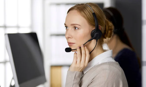 Femme d'affaires amicale parlant par casque pendant qu'elle est assise au bureau. Centre d'appels et divers groupes de personnes en affaires — Photo