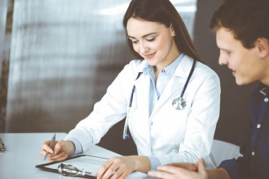 Dost canlısı genç bayan-doktor, birlikte bir dolapta masada otururken, hastasını dinliyor. Hekim elinde ilaç doldurmak için bir dosya tutuyor.