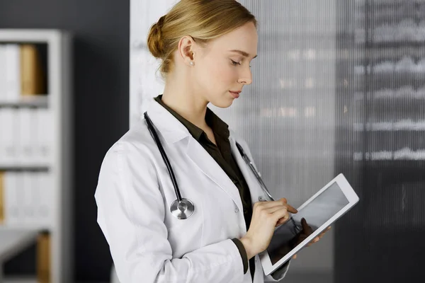 Friendly smiling female doctor using clipboard in clinic. Portrait of friendly physician woman at work place. Perfect medical service in hospital