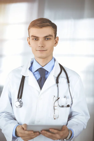 El joven doctor sonríe alegremente a la cámara, mientras está de pie en una soleada oficina del hospital, sosteniendo una tableta de computadora en sus manos. Servicio médico perfecto en la clínica. Feliz futuro en la medicina y —  Fotos de Stock