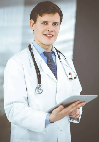 Ein junger freundlicher Arzt checkt ein paar Informationen auf seinem Computer-Tablet. Porträt eines professionellen Arztes bei der Arbeit in einer Klinik. Medizinkonzept — Stockfoto