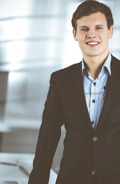 Empresário bem sucedido amigável em um terno preto está de pé em seu local de trabalho. Retrato de um jovem especialista em um escritório moderno. Conceito de negócio — Fotografia de Stock