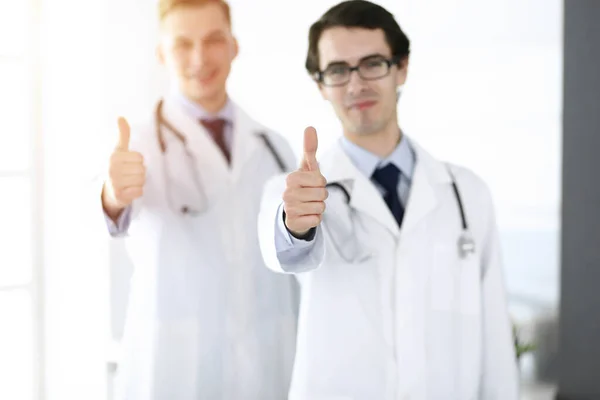 Twee mannelijke artsen staan als een team en tonen duimen omhoog als een symbool van de beste service voor patiënten in zonnige kliniek. Geneesmiddelenconcept — Stockfoto