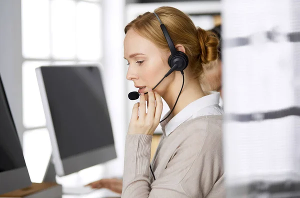 Femme d'affaires amicale parlant par casque pendant qu'elle est assise au bureau. Centre d'appels et divers groupes de personnes en affaires — Photo