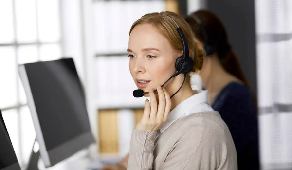 Femme d'affaires amicale parlant par casque pendant qu'elle est assise au bureau. Centre d'appels et divers groupes de personnes en affaires — Photo