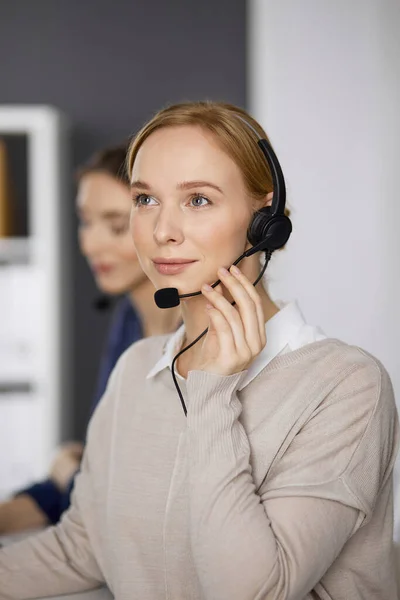 Femme d'affaires amicale parlant par casque pendant qu'elle est assise au bureau. Centre d'appels et divers groupes de personnes en affaires — Photo