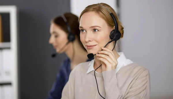 Femme d'affaires amicale parlant par casque pendant qu'elle est assise au bureau. Centre d'appels et divers groupes de personnes en affaires — Photo