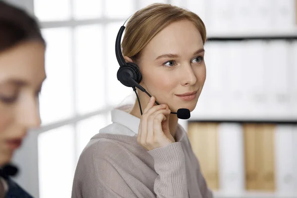 Femme d'affaires amicale parlant par casque pendant qu'elle est assise au bureau. Centre d'appels et divers groupes de personnes en affaires — Photo