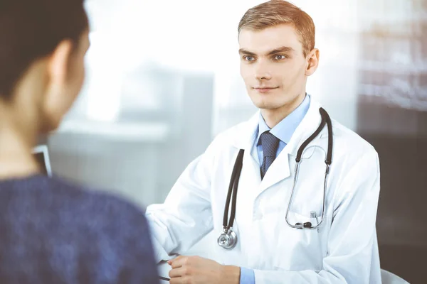 Doctor man, with a stethoscope, is listening to his patient, while sitting at the desk in the sunny cabinet at hospital. Perfect medical service in clinic. Happy future in medicine and healthcare