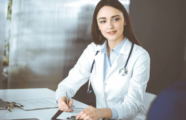 Sorrindo jovem mulher-médico está enchendo recomendações médicas usando uma área de transferência. Terapeuta no trabalho em um escritório de clínica. Conceito de medicina — Fotografia de Stock