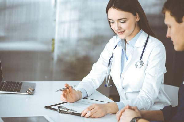 Friendly young woman-doctor is listening to her patient, while they are sitting together at the desk in a cabinet. Physician is holding a clipboard in her hands for filling up medication