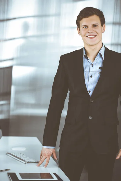 Un homme d'affaires sympathique et prospère en costume noir se tient debout sur son lieu de travail. Portrait d'un jeune spécialiste dans un bureau moderne. Concept d'entreprise — Photo