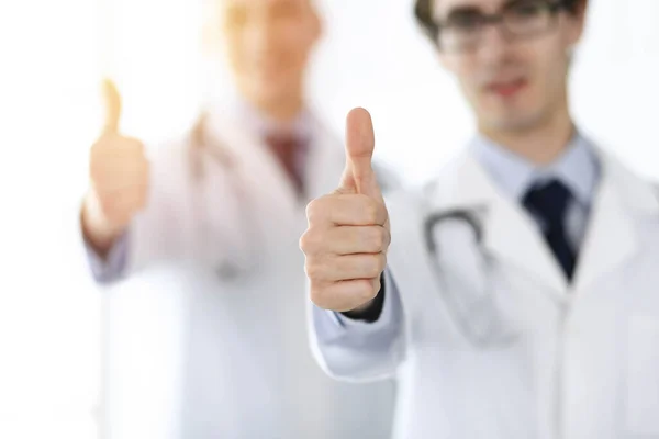 Dois médicos do sexo masculino em pé como uma equipe e mostrando os polegares para cima como um símbolo do melhor serviço para pacientes em clínica ensolarada. Conceito de medicina — Fotografia de Stock