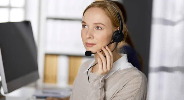 Femme d'affaires amicale parlant par casque pendant qu'elle est assise au bureau. Centre d'appels et divers groupes de personnes en affaires — Photo