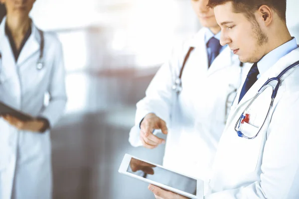 Een groep artsen controleert medische namen op een tablet, met een verpleegkundige met een klembord op de achtergrond, terwijl ze samen staan in een zonnig ziekenhuiskantoor. Artsen klaar om te onderzoeken en — Stockfoto