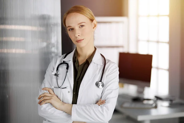 Friendly smiling female doctor standing in sunny clinic. Portrait of friendly physician woman. Medicine concept