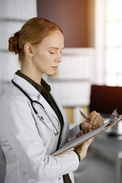 Friendly smiling female doctor using clipboard in sunny clinic. Portrait of friendly physician woman at work. Medicine concept