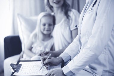 Doktor ve hasta. Pediyatrist, annesiyle birlikte küçük kızı muayene ederken pano kullanıyor. Sağlık muayenesinde mutlu beyaz çocuk. Tıp konsepti