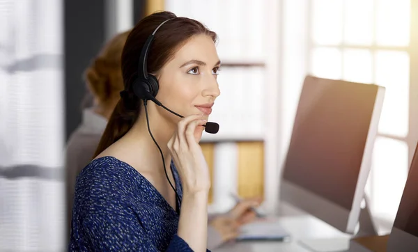 Femme d'affaires asiatique ou latino-américaine parlant par casque dans un bureau ensoleillé. Centre d'appels et divers groupes de personnes en affaires — Photo