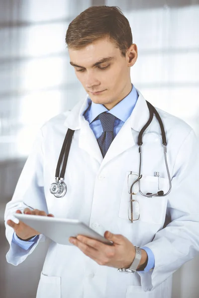 Νεαρός γιατρός στέκεται σε ένα γραφείο νοσοκομείου, κρατώντας μια ταμπλέτα υπολογιστή στα χέρια του. Τέλεια ιατρική υπηρεσία στην κλινική. Ευτυχές μέλλον στην ιατρική και την υγειονομική περίθαλψη — Φωτογραφία Αρχείου