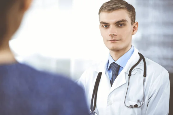 El doctor, con un estetoscopio, está escuchando a su paciente, mientras está sentado en el escritorio del gabinete del hospital. Servicio médico perfecto en la clínica. Futuro feliz en medicina y salud —  Fotos de Stock