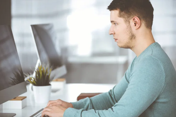 Jeune homme d'affaires confiant, portant un pull vert, est assis sur le bureau dans un bureau moderne, tout en travaillant sur l'ordinateur sur son nouveau projet. Headshot de l'entrepreneur masculin ou it-spécialiste à — Photo