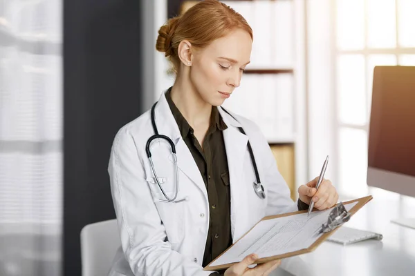 Friendly smiling female doctor using clipboard in sunny clinic. Portrait of friendly physician woman at work place. Perfect medical service in hospital. Medicine concept