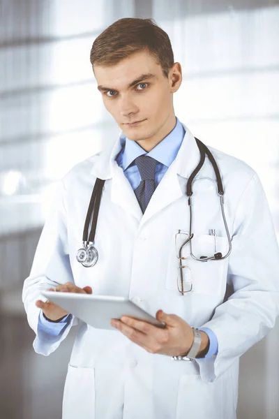 Fiatal orvos férfi áll egy kórházi irodában, kezében egy számítógépes táblával. Tökéletes orvosi ellátás a klinikán. Boldog jövőt az orvostudományban és az egészségügyben — Stock Fotó