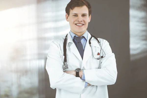 Un giovane dottore positivo sta sorridendo alla telecamera. Ritratto di medico professionista in clinica soleggiata — Foto Stock