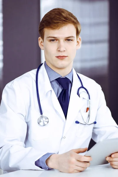 Blonde männliche Arzt sitzt und arbeitet mit Tablet-Computer in der Klinik an seinem Arbeitsplatz, Nahaufnahme. Junger Arzt bei der Arbeit. Perfekter medizinischer Service, medizinisches Konzept — Stockfoto