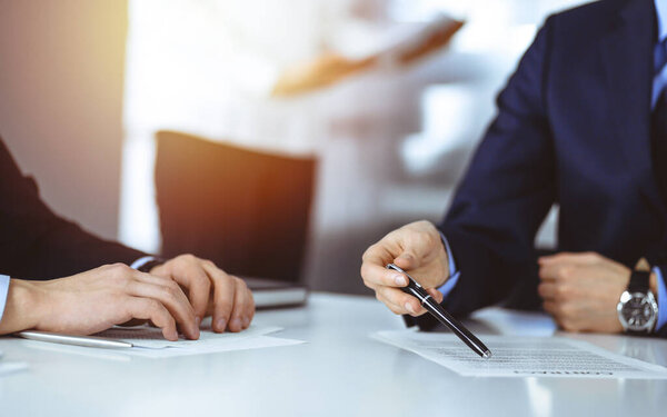 Business people discussing a contract at meeting or negotiation, sitting at the desk together, and an unknown woman on the background, close-up. Group of unknown businessmen in a sunny modern office