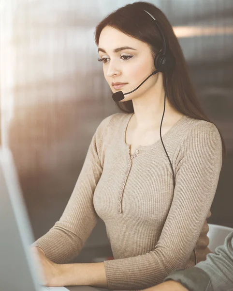 Jeune fille amicale dans les écouteurs parle à un client des entreprises, tout en étant assis au bureau ensoleillé. Opérateurs de centre d'appels au travail — Photo